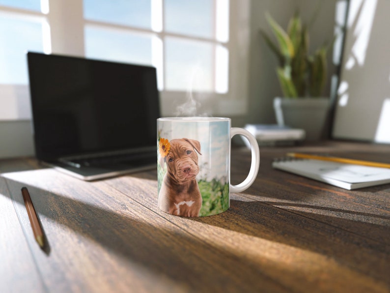 May include: A white ceramic mug with a brown puppy with a yellow flower behind its ear printed on it. The puppy is sitting in a grassy field.