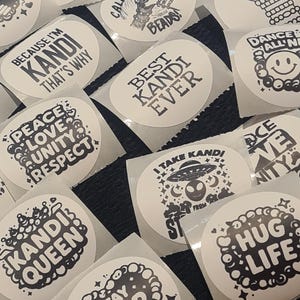 May include: A selection of white, oval stickers with black text and graphics. The stickers display phrases such as "Best Kandi Ever," "Kandi Queen," and "Stay PLUR," alongside various designs, including a smiley face and an alien.