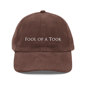 May include: A brown corduroy baseball cap with white embroidery that reads "FOOL OF A TOOK".