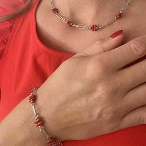 Peut inclure: Un ensemble collier et bracelet en argent avec des perles de corail rouge. Le collier et le bracelet ont un design à maillons de chaîne. Les bijoux sont présentés sur un fond rouge.