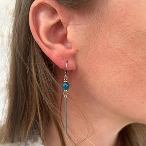 May include: A silver earring with a teal bead and a long, rectangular silver bar. The earring is hanging from a silver hoop. The earring is worn on the ear.