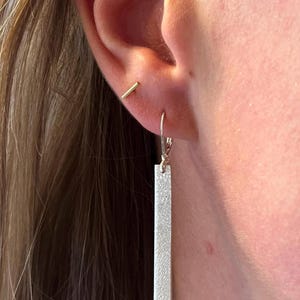 Peut inclure: Gros plan sur une oreille ornée de deux boucles d'oreilles. L'une est une petite barre dorée. L'autre est une longue boucle d'oreille rectangulaire argentée avec une surface texturée et une fermeture à levier. Les boucles d'oreilles sont sur la peau et les cheveux.