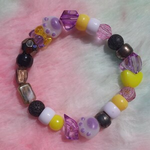 May include: A stretch bracelet featuring a variety of beads including white and yellow round plastic beads, purple paw print shaped beads, translucent purple faceted beads, black porous lava beads, and metallic bronze rectangular and round beads.
