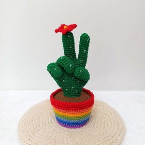 May include: A green crocheted cactus shaped like a peace sign with a red flower on top, in a rainbow striped pot, sitting on a beige crocheted mat.