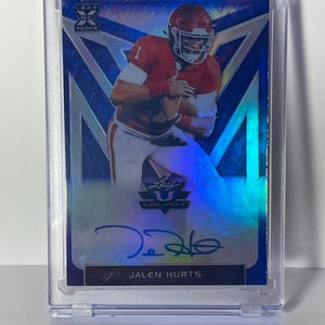 以下が含まれることがあります： 保護ケースに入ったJalen Hurtsのルーキーフットボールカード。カードには、赤と白のユニフォームを着た選手が描かれており、下部にはサインと「Jalen Hurts」の名前があります。カードは青と銀のデザインです。