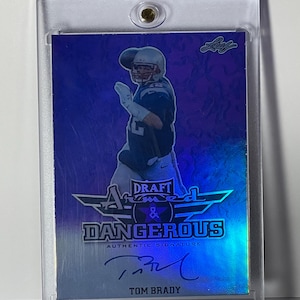 Puede incluir: Una tarjeta coleccionable de Tom Brady en una funda protectora. La tarjeta presenta una foto de Brady con uniforme de f&uacute;tbol, con las palabras "Draft Armed & Dangerous" y "Authentic Signature" impresas. La tarjeta tiene un esquema de color azul y plateado.
