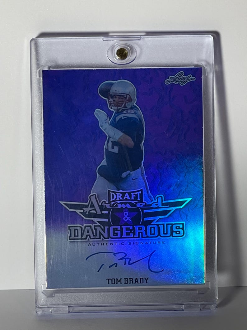 Puede incluir: Una tarjeta coleccionable de Tom Brady en una funda protectora. La tarjeta presenta una foto de Brady con uniforme de f&uacute;tbol, con las palabras "Draft Armed & Dangerous" y "Authentic Signature" impresas. La tarjeta tiene un esquema de color azul y plateado.