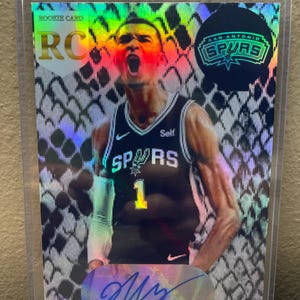 Könnte beinhalten: Eine Rookie-Karte der San Antonio Spurs mit einem Spieler in einem schwarz-weißen Trikot mit der Nummer 1. Die Karte hat einen holografischen Hintergrund und die Unterschrift des Spielers. Die Karte ist in einer Schutzhülle.