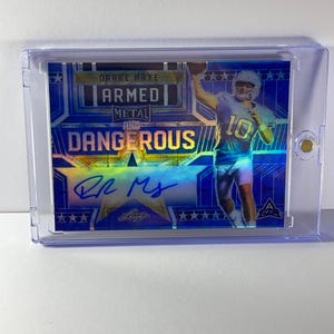 Könnte beinhalten: Eine blaue und silberne Sport-Sammelkarte mit einem Footballspieler, der einen Ball wirft. Die Karte trägt den Text "ARMED METAL AND DANGEROUS" und die Unterschrift des Spielers. Die Karte ist in einem transparenten Kunststoffhalter eingeschlossen.