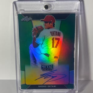 May include: A baseball card featuring Shohei Ohtani in a clear protective case. The card has a holographic design with the words "METAL DRAFT" and "SHOHEI OHTANI" printed on it. Ohtani is wearing a red cap and jersey with the number 17.