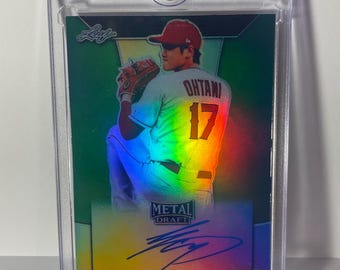 May include: A baseball card of Shohei Ohtani in a clear protective case. The card has a holographic design with the words "METAL DRAFT" and "SHOHEI OHTANI" printed on it. Ohtani is wearing a red cap and jersey with the number 17.