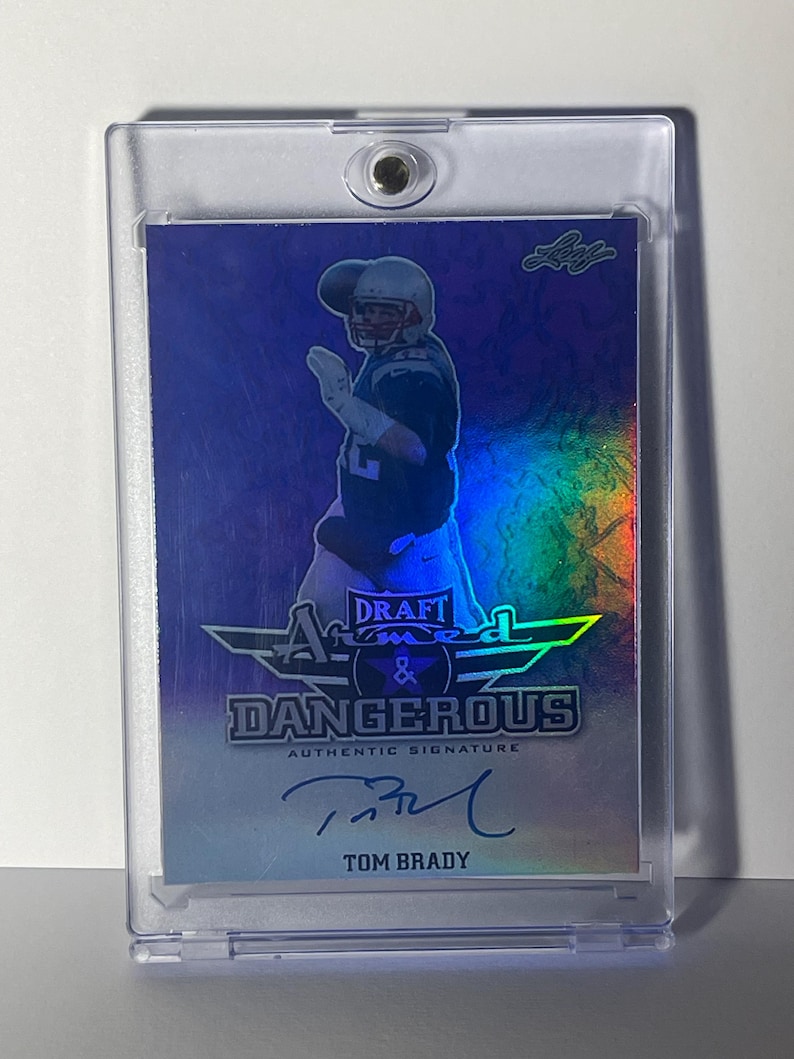 Puede incluir: Una tarjeta coleccionable de Tom Brady "Draft & Dangerous" en un estuche protector. La tarjeta presenta una imagen de Brady con su uniforme de f&uacute;tbol, con su firma debajo del texto. La tarjeta tiene un fondo hologr&aacute;fico.