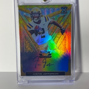 以下が含まれることがあります： 白と紫のユニフォームを着たフットボール選手が描かれたスポーツトレーディングカード。カードは金と虹色のデザインで、選手のサインと「JUSTIN JEFFERSON」の名前が記されています。カードは透明な保護ホルダーに入っています。