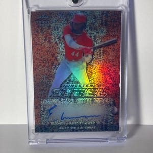 May include: A baseball trading card featuring Elly De La Cruz in a red uniform, swinging a bat. The card has a holographic background with the words "Tungsten Steel" and an autograph. The card is encased in a clear protective holder.
