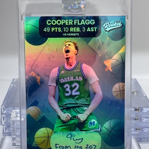 May include: A Cooper Flagg rookie basketball card in a protective case. The card features a player in a green Dallas Mavericks jersey with the number 32. Text includes "49 PTS, 10 REB, 3 AST" and "From the 207."