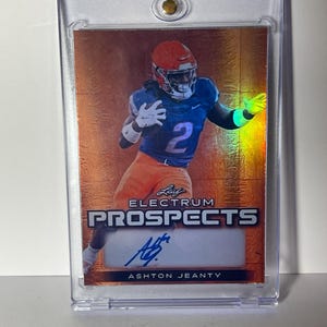 May include: A football trading card featuring Ashton Jeanty in a blue and orange uniform. The card is encased in a clear protective holder. The card has the text "Leaf Electrum Prospects" and an autograph.