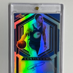 Puede incluir: Tarjeta de baloncesto de Stephen Curry en una funda protectora. La tarjeta muestra a Curry con uniforme azul y dorado, sosteniendo un balón de baloncesto. La palabra "CONTINUUM" y la firma de Curry están presentes.