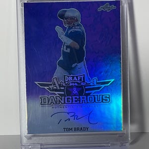 Puede incluir: Una tarjeta de f&uacute;tbol de Tom Brady en una funda protectora. La tarjeta presenta una imagen del jugador, el texto "DRAFT ARMED & DANGEROUS AUTHENTIC SIGNATURE" y la firma de Brady. La tarjeta tiene un esquema de color azul y plateado.