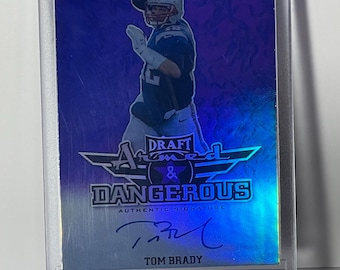 May include: A Tom Brady football card in a protective case. The card features a player image, the text "DRAFT ARMED & DANGEROUS AUTHENTIC SIGNATURE", and Brady's signature. The card has a blue and silver color scheme.