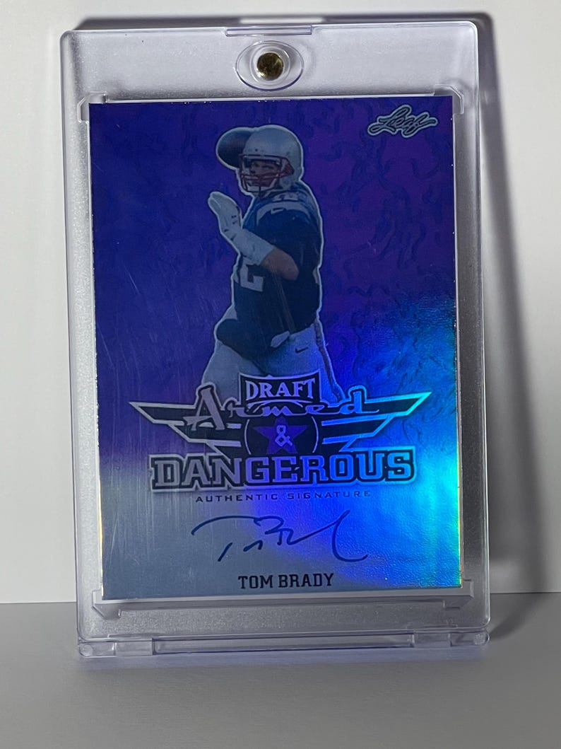 Puede incluir: Una tarjeta de f&uacute;tbol de Tom Brady en una funda protectora. La tarjeta presenta una imagen del jugador, el texto "DRAFT ARMED & DANGEROUS AUTHENTIC SIGNATURE" y la firma de Brady. La tarjeta tiene un esquema de color azul y plateado.