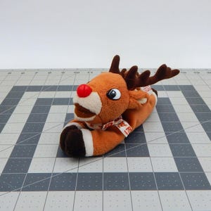 Könnte beinhalten: Ein Plüsch-Rentier-Spielzeug mit roter Nase, braunem Geweih und einem rot-weiß karierten Schal. Das Rentier liegt auf einer grau-weiß karierten Oberfläche. Das Spielzeug hat einen weißen Bauch und braune Hufe.