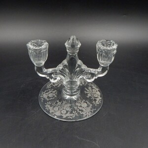 May include: Clear glass candelabra with two arms and a central finial. The base features an etched floral design. The candelabra is designed to hold two candles. The overall design is ornate and decorative.