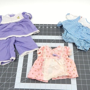 May include: Three doll outfits are displayed. One is a lavender top and shorts set with white lace trim. Another is a blue and white gingham dress. The third is a pink floral dress with lace trim.