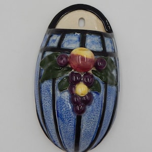 May include: Blue ceramic wall pocket with a grape and apple design. The oval-shaped pocket has a blue background with black vertical stripes. A cluster of purple grapes, a red and yellow apple, and green leaves are featured.