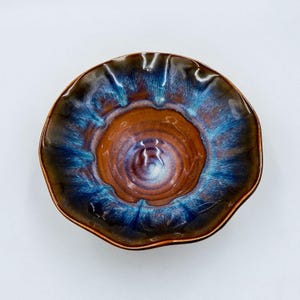 Puede incluir: Un cuenco de cerámica hecho a mano con una forma única y orgánica. El cuenco presenta una rica paleta de colores marrón, azul y bronce, con un acabado brillante. El borde tiene un borde ligeramente ondulado, lo que aumenta su atractivo artístico. Este cuenco decorativo es ideal para servir o exhibir.