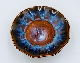 May include: A handmade ceramic bowl with a unique, organic shape. The bowl features a rich color palette of brown, blue, and bronze, with a glossy finish. The rim has a slightly ruffled edge, adding to its artistic appeal. This decorative bowl is ideal for serving or display.