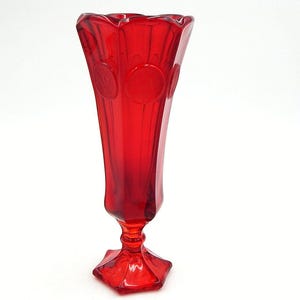 Puede incluir: Un jarrón de cristal rojo vibrante con una parte superior acampanada y una base de pedestal. El jarrón presenta una serie de crestas verticales y elementos decorativos circulares. El diseño elegante es adecuado para exhibir flores o como acento decorativo.