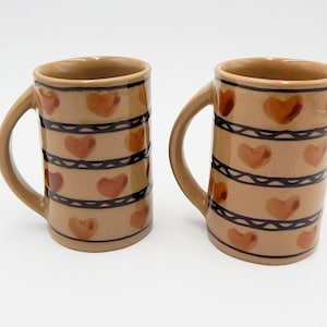 Puede incluir: Dos tazas de cerámica con base de color beige. Cada taza presenta un patrón repetido de corazones marrones y líneas negras en zigzag. Las tazas tienen un asa curva y una forma cilíndrica.