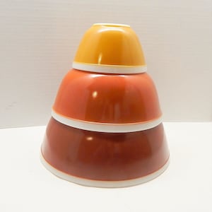 May include: Stacked set of three vintage mixing bowls in shades of orange and yellow. The bowls are nested, with a white rim on each. The top bowl is a bright yellow, the middle bowl is orange, and the bottom bowl is a deep reddish-orange.