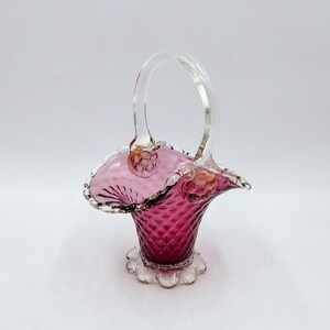 May include: A deep magenta glass basket featuring a diamond-patterned body, a clear arched handle, and a ruffled rim. The base is decorated with a series of clear glass spheres, and the handle attaches to the body with two clear floral accents.