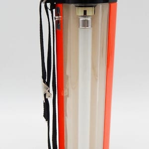 Könnte beinhalten: Eine tragbare Leuchtstofflampe mit einem schwarzen und orangefarbenen Kunststoffgehäuse. Sie hat eine transparente Kunststoffabdeckung und eine weiße Leuchtstoffröhre. Die Lampe hat einen schwarzen Trageriemen.