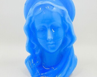May include: A blue candle shaped like a woman's face with a halo. The candle has a smooth, glossy finish and a wick at the top. The face has closed eyes and flowing hair. The candle is a solid, vibrant blue color.