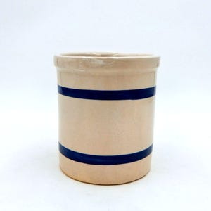 May include: A beige ceramic utensil holder with two dark blue horizontal stripes. The cylindrical container has a slightly flared rim and a smooth, glossy finish. This kitchenware item is suitable for storing cooking tools.