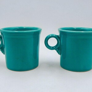 May include: Two turquoise ceramic mugs with rounded handles. The mugs have a simple, classic design with a slightly flared rim and a smooth, glossy finish. The handles are circular and attached to the side of the mug.
