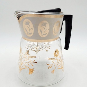 May include: A clear glass carafe featuring a wide band at the top with gold oval floral motifs. The lower section is decorated with gold wheat and flower patterns. It includes a black plastic handle and a black plastic lid attached at the top.