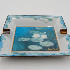 Op de afbeelding: Vierkant porseleinen asbak met een blauw en wit waterlelie ontwerp. De asbak heeft een gouden rand en de handtekening van de kunstenaar "Claude Monet".