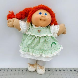 Può includere: Una bambola vintage Cabbage Patch Kids con codini di filo rosso, occhi blu e un viso sorridente. La bambola indossa un vestito verde floreale con finiture in pizzo bianco, leggings bianchi e scarpe color crema. Il logo Cabbage Patch Kids è sul vestito.