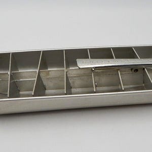 May include: A vintage aluminum ice cube tray with a handle and a lever for releasing cubes. The tray is rectangular with dividers creating individual cube compartments. The metal has a brushed finish.