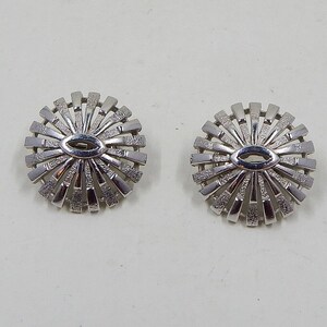 May include: A pair of silver-coloured, circular clip-on earrings. Each earring has a central oval shape surrounded by radiating, textured bars, creating a sunburst design. The earrings are set against a plain white background.