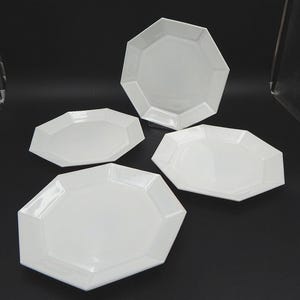 May include: Four white, geometric-shaped ceramic plates. The plates have a glossy finish and a unique, faceted design. The plates are arranged on a dark surface, showcasing their clean lines and modern aesthetic. Ideal for serving food.