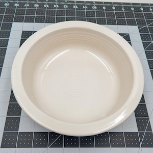 May include: A cream-colored, round ceramic bowl with a wide rim. The bowl has a shallow depth and a smooth, glossy finish. The interior features concentric circles at the center. Ideal for serving soups, salads, or side dishes.
