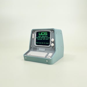 Op de afbeelding: Een teal-kleurige retrocomputer met een scherm met "UOS" en andere tekst. De computer heeft een toetsenbord, twee witte knoppen en een kleine luidspreker. De tekst op het scherm luidt "ROBCO INDUSTRIES UNIFIED OPEATING SYSTEM Copyright 2075-2077 Press any key to start."