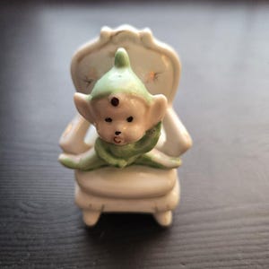 Puede incluir: Pequeña figura vintage de porcelana de un elfo sentado en una silla color crema. El elfo tiene un gorro y ropa verdes, con un tono de piel claro y tres pequeños puntos marrones en la frente. La silla tiene respaldo curvo y delicados detalles florales.