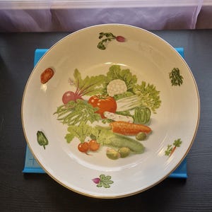 Könnte beinhalten: Eine weiße Keramikschüssel mit goldfarbenem Rand zeigt ein farbenfrohes Gemüse-Design. Die Schüssel ist mit verschiedenen Gemüsesorten gefüllt, darunter Karotten, Tomaten, Paprika und Zwiebeln.