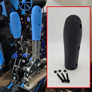 May include: A black shifter knob with a textured surface and two holes on the side. The knob is sitting on a white surface with four black screws and nuts.