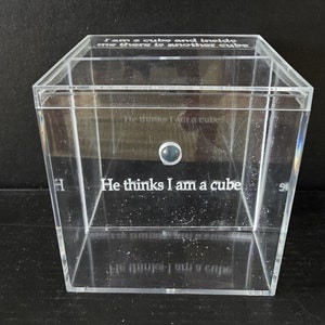 Può includere: Una scatola in acrilico trasparente con il testo "Sono un cubo e dentro di me c'è un altro cubo" sulla parte superiore e "Lui pensa che io sia un cubo" sulla parte anteriore. La scatola ha un piccolo pulsante blu sulla parte anteriore.
